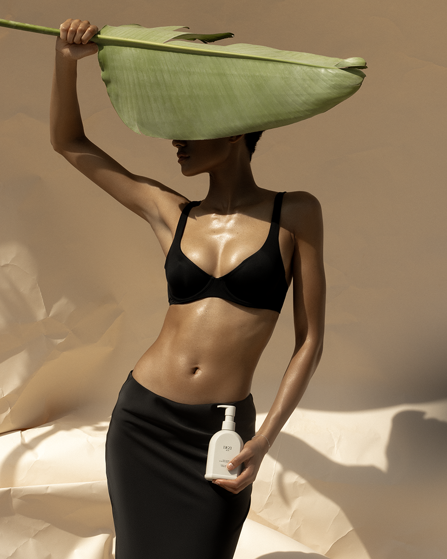 a woman holding a banana leaf and the body gloss body lotion bottle. the leaf is shading her face. her body looks very hydrated and is glistening in the sun. 