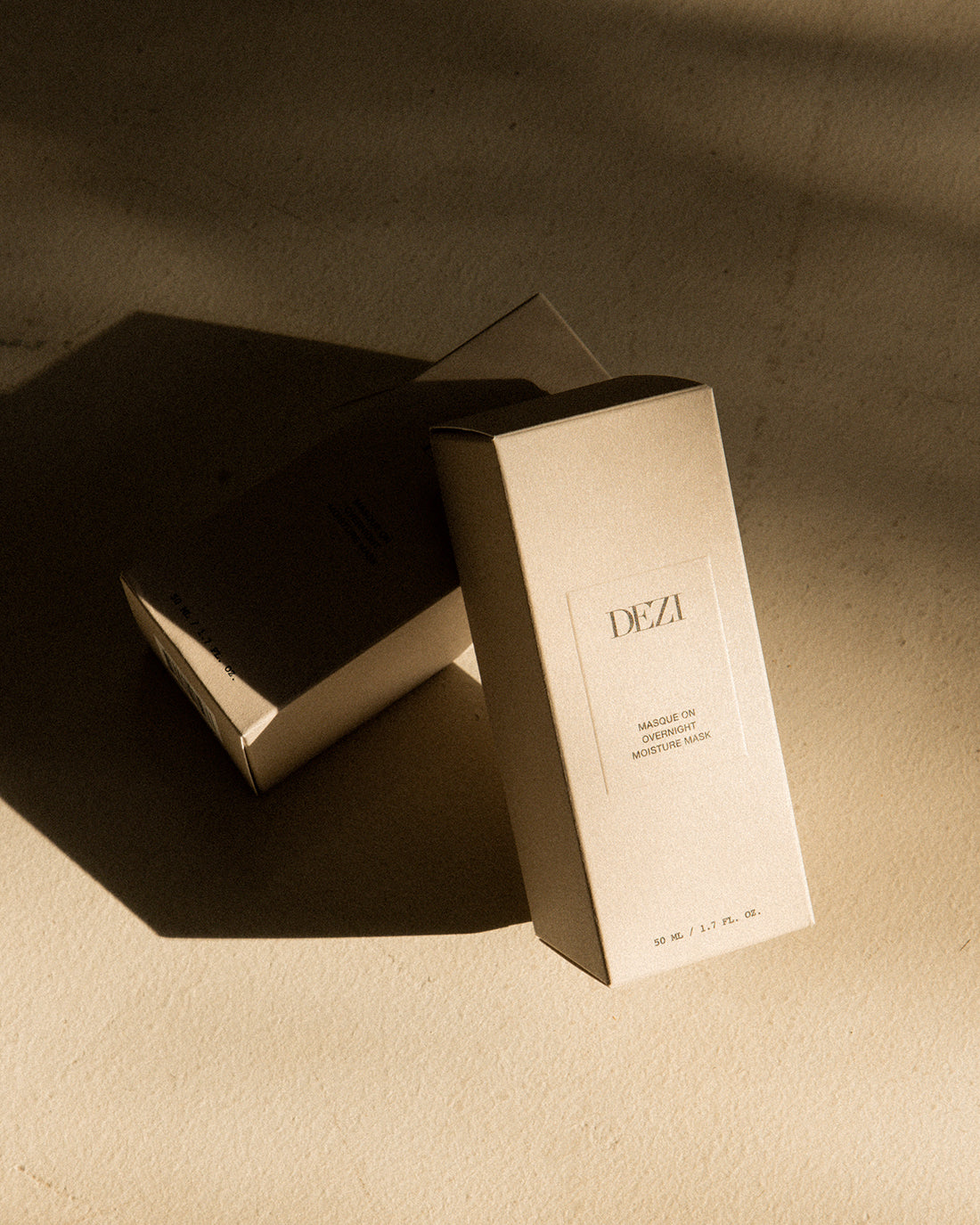 this shows two dezi skin moisture mask boxes leaning on each other. the sunlight is hitting them, casting long dark shadows on the stone floor. 